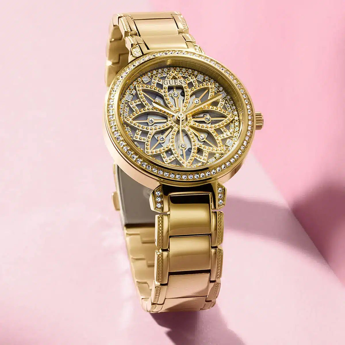 Guess Lily watch worn on wrist with gold case and bracelet against pink background